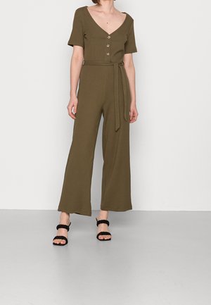 Combinaison côtelée vert olive avec un décolleté en V, des manches courtes, des détails de boutons et une taille à nouer, associée à des sandales noires à lanières.