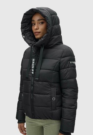 Femme portant une veste matelassée noire à capuche avec fermeture éclair devant et poches latérales, debout devant un fond clair uni.