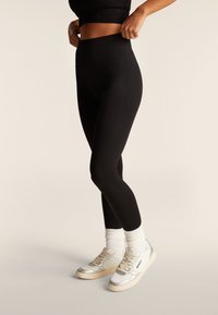 Donna che indossa leggings neri a vita alta, che tira la cintura, abbinati a calzini bianchi e sneakers bianche su sfondo beige.