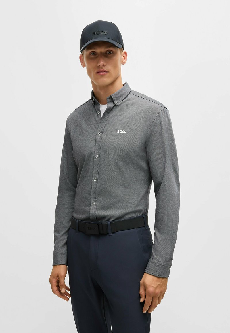 Grå långärmad skjorta med knappar och texturerad tyg, samt en vit logotyp. Kombinerad med mörka byxor och en svart keps med matchande logotyp.