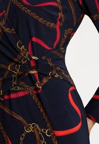 Tmavomodré šaty s potlačou červeno-hnedých spletených lán a opasku, s detailom zlatej okrúhlej pracky v páse.