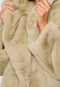 Des mains serrant le col d'un manteau en fausse fourrure beige et duveteux, mettant en valeur la texture et la chaleur du vêtement.