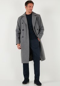 Grauer Woll-Peacoat mit doppelreihigen Knöpfen, Reverskragen und seitlichen Taschen, kombiniert mit dunkler Hose und dunklen Stiefeletten.