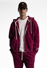 Sweat à capuche bordeaux zippé avec cordons blancs, poches kangourou et coupe slim. Associé à un pantalon de jogging bordeaux assorti et une casquette marron.