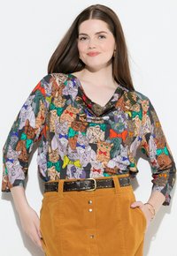 Colorful, patterned long-sleeve top featuring cats in various colors and bow ties, with a draped neckline, paired with a tan corduroy skirt.
