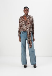 Leopardenmuster-Wickeloberteil aus Satin mit Bindeschluss, kombiniert mit hellblauen Schlagjeans mit Patches und schwarzen spitz zulaufenden Ballerinas.