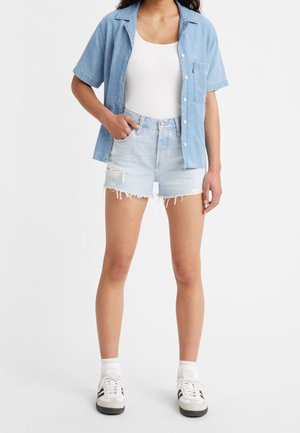 Camisa de mezclilla azul clara, camiseta blanca sin mangas y pantalones cortos desgastados de mezclilla azul clara. Zapatillas blancas con detalles en negro. Conjunto casual con un ajuste relajado.