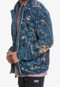 Chaqueta de forro polar azul con patrones naranjas, con cremallera en el frente, puños elásticos y un bolsillo en el pecho de un material contrastante.
