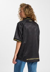 Μαύρη κοντομάνικη μπλούζα με γεωμετρικό σχέδιο, κίτρινες λεπτομέρειες στον γιακά και στα μανίκια, και κείμενο "ΔΙΕΘΝΗΣ" στο στρίφωμα.