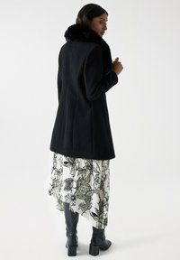 Manteau noir en laine avec un col en fausse fourrure, associé à une jupe blanche à motifs floraux et des bottes noires jusqu'aux genoux. Vue de dos, texture lisse.