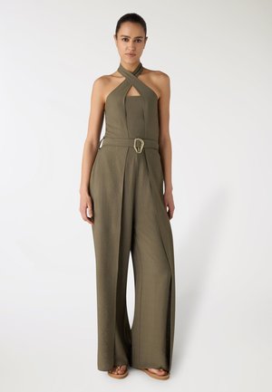 Donna che indossa una tuta pantalone verde oliva a collo halter con gambe larghe e cintura abbinata con fibbia dorata, in piedi contro uno sfondo semplice.