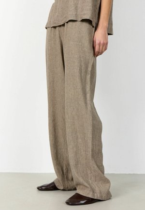 Osoba nosí voľné svetlohnedé ľanové nohavice a zladený top, doplnené tmavohnedými špicatými topánkami na svetlej podlahe a bielom pozadí.