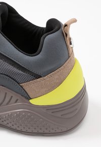 Baskets gris et marron avec une tige texturée, un accent jaune au talon et un col rembourré. Elles possèdent une semelle en caoutchouc avec un motif de bande de roulement.