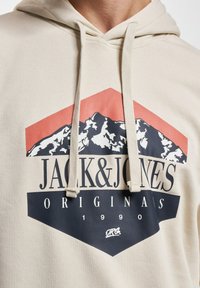 Beige Kapuzenpullover mit Kordelzug. Beinhaltet ein grafisches Design mit Bergen, "JACK&JONES"-Text und kontrastierenden dunkelblauen sowie korallenfarbenen Akzenten.