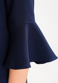 Mörkblå klänningärm med en klockformad design, tillverkad av ett lent tyg med en subtil lyster och rena sömmar längs fållen.