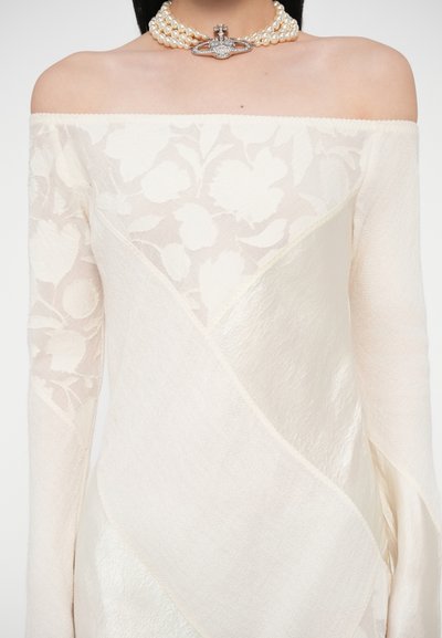 Šaty s odhalenými ramenami v svetlo krémovej farbe s kvetinovou čipkovanou vrstvou, s textúrovanou látkou a diagonálnymi panelmi. Náhrdelník z korálkových perál s brošňovým doplnkom.