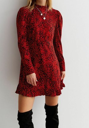 Woman wearing a red, long-sleeve, high-neck dress with black leopard spots and ruffled hem paired with black knee-high boots.