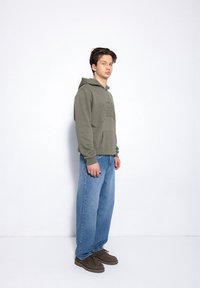 Oliwkowy hoodie z kieszenią z przodu i wytłoczonym napisem, zestawiony z jasnoniebieskimi, luźnymi dżinsami i brązowymi butami.