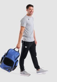Camiseta gris deportiva y pantalones de chándal negros, combinados con zapatillas deportivas grises. La mochila azul tiene detalles de malla y un logotipo visible.