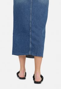 Donkerblauwe denim midi-jurk met een ruwe zoom, voorzien van een centrale achternaad. Gecombineerd met zwarte platte sandalen.