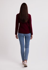 Una camiseta de manga larga burdeos combinada con jeans ajustados de color azul claro y zapatillas blancas y rosas, vista desde atrás contra un fondo liso.