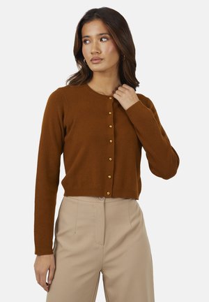 Kobieta ubrana w brązowy sweter na guziki i beżowe spodnie z wysokim stanem, patrząca w lewo z ręką przy kołnierzu.