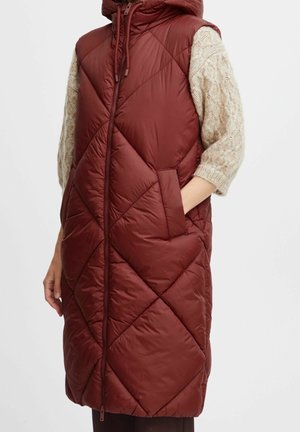 Ærmeløs, vatteret, burgunderrød hættetrøje med lynlås foran, båret over en beige kabelstrikket sweater med albuelange ærmer.