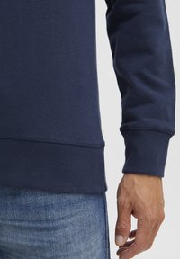 Jūras spēku džemperis ar ribotu piedurkņu un apakšmali, apvienots ar zilo džinsingu. Materiāls izskatās mīksts un tam ir gluda tekstūra.