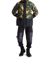 Kamouflagejacka i grönt och orange med svarta detaljer, tillsammans med svarta sweatpants med vadderade svarta paneler och svarta stövlar.