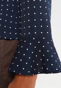 Tessuto blu navy con pois bianchi, maniche svasate, indossato sopra pantaloni marroni su un avambraccio visibile.