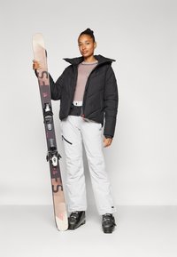 Tenue de ski comprenant une veste matelassée noire, un pantalon gris et un pull à motifs. Tenant un ski avec des fixations noires et un logo de marque.