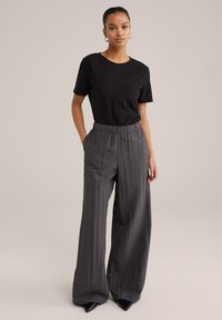 Melnas acu adīšanas T-krekls apvienots ar brīvi pieguļošām, pelēkām platām biksēm ar elastīgu jostasvietu un gludu auduma tekstūru. Melnas norādītu purngalu kurpes.