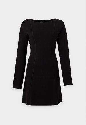 Femme portant une mini-robe noire à manches longues, des bottes marron mi-mollet, un collier en chaîne dorée, tenant un grand sac noir dans sa main gauche.