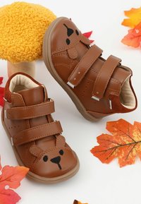 Chaussures marron pour tout-petits avec un motif visage d'ours et double fermeture Velcro, entourées de feuilles d’automne et d’un champignon décoratif jaune.