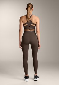 Reggiseno sportivo marrone con design a taglio e leggings a vita alta coordinati. Il tessuto è liscio, elastico, con un marchio minimale e calzature nere.