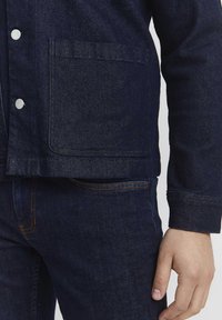 Denim jacket in dark blue with visible stitching, silver snap buttons, and a front pocket. Paired with matching dark blue jeans.