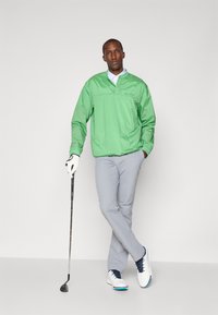 Lichtgroene waterdichte jas met een halve rits, grijze chinos en witte golfschoenen met blauwe accenten, vergezeld van een golfclub en handschoen.
