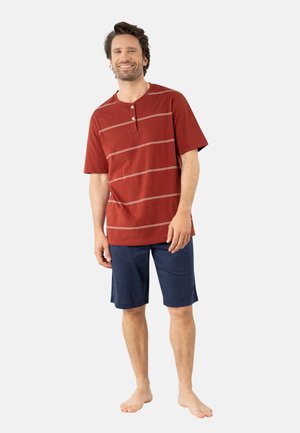 Homme souriant debout pieds nus, portant une chemise rouge à manches courtes avec de fines rayures blanches et un short bleu marine arrivant aux genoux.