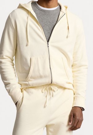Man wearing cream zip-up hoodie and matching drawstring sweatpants over a grey crew neck sweater, hands in pockets, showing wristwatch.