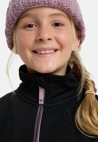 Černá fleecová bunda s vysokým límcem a růžovým zipem. Osoba má na sobě pletenou světle fialovou čepici a vykazuje uvolněný výraz.