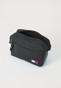 Čierna messenger taška s matným povrchom, s uzatvorením na zips a nastaviteľným popruhom. Má farebný logový nášivku a šedé akcenty.
