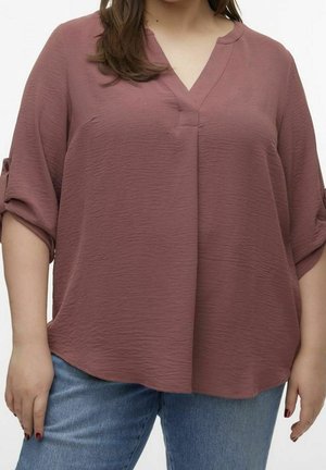 Mujer con blusa texturizada color malva con cuello en V y mangas remangadas, y pantalones vaqueros azules, de pie con las manos relajadas a los lados.