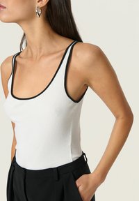 Weißes, geripptes Tanktop mit schwarzem Saum, das über einen Rundhalsausschnitt und dünne Träger verfügt, kombiniert mit hochtaillierten schwarzen Hosen.