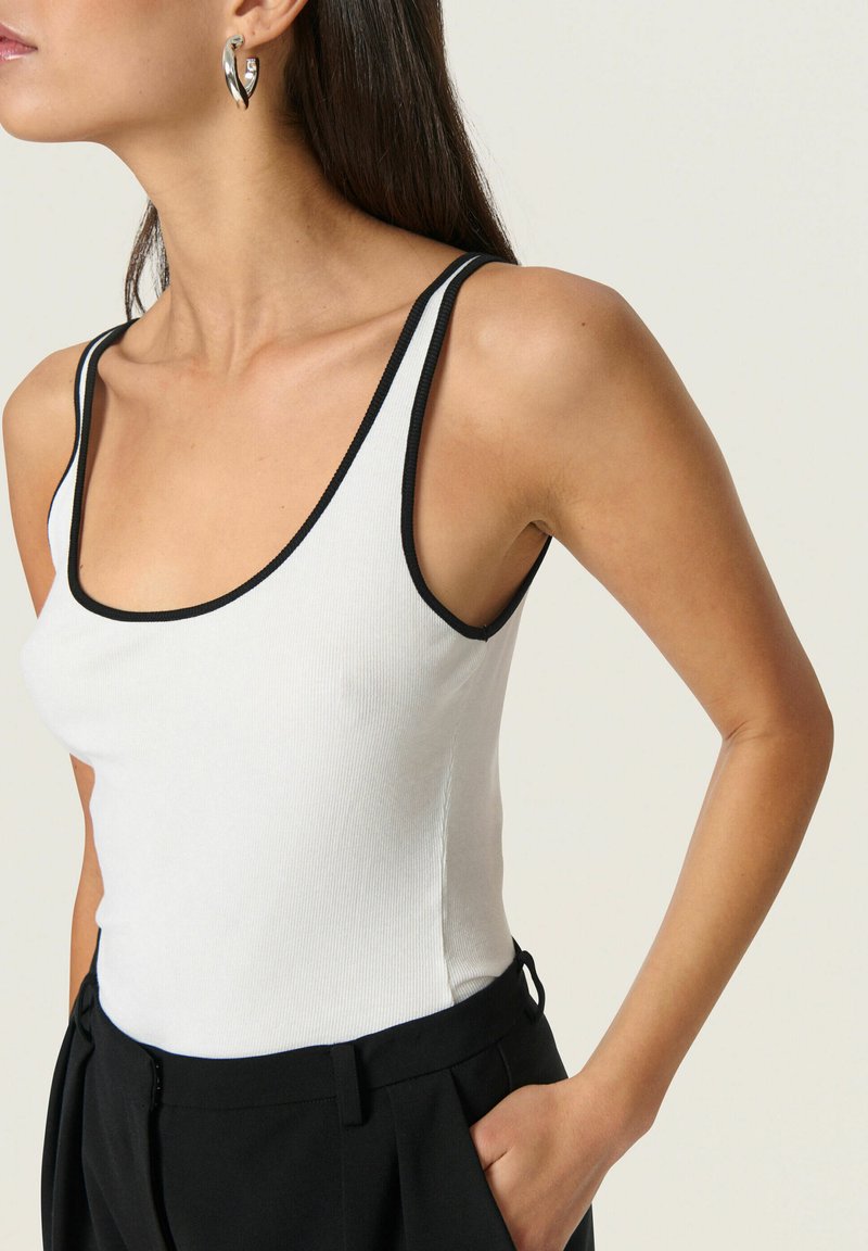 Top de tank blanco con ribetes negros, con escote redondeado y tirantes finos, combinado con pantalones negros de cintura alta.