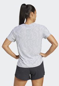Camisa deportiva de manga corta en gris claro con un patrón de ondas texturizado, combinada con pantalones cortos negros. La vista trasera muestra un diseño y ajuste sutiles.