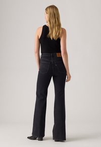 Vrouw met lang blond haar, gekleed in een zwart mouwloos top, hoge donkere jeans met achterzakken en zwarte haklaarzen, staande met haar rug naar ons toe.