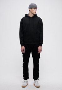 Crna dukserica s kapuljačom i vezicama, prednji džep, u kombinaciji s crnim cargo hlačama i bijelim tenisicama. Sivi pleteni beanie dovršava odjevnu kombinaciju.