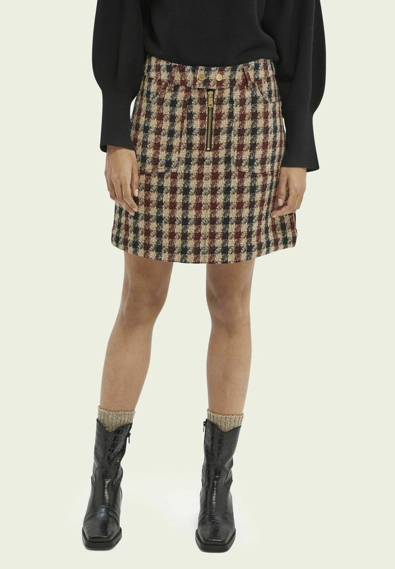 Femme portant une jupe à carreaux avec une fermeture éclair à l'avant et des poches, un haut noir à manches longues, des chaussettes beiges et des bottines noires.