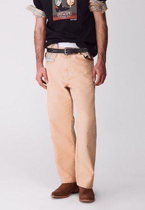 Mężczyzna w beżowych spodniach, czarnym pasku, czarnej koszulce z grafiką, podwiniętymi rękawami koszuli w kratę oraz brązowych zamszowych butach na jednolitym tle.