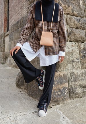 Personne portant une veste à motif pied-de-poule, une chemise blanche, un pantalon large noir, des baskets multicolores et un petit sac bandoulière beige, debout devant un mur de pierre.